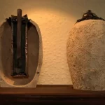 Imagem conceitual mostrando a Bateria de Bagdá conectada a um painel metálico antigo, com inscrições cuneiformes e luzes simbólicas, sugerindo seu uso como fonte energética para dispositivos védicos.